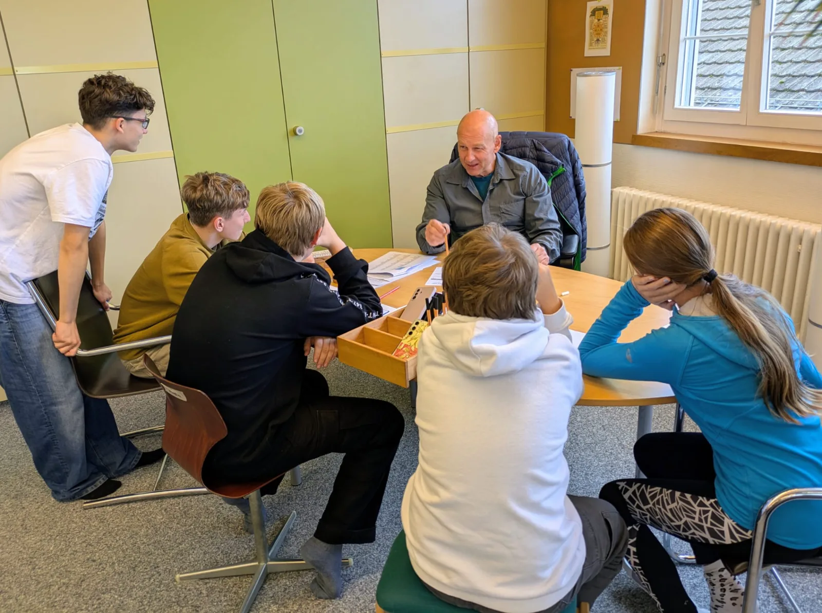 Lehrperson am Tisch erklärt etwas einer Gruppe von Jugendlichen